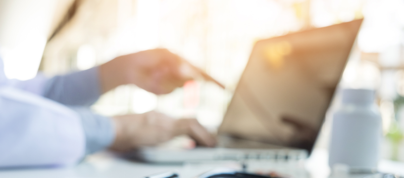 A blurred image shows a person using a laptop, with one hand on the keyboard and the other pointing at the screen—perhaps reviewing a patient safety structural measure. A bottle and eyeglasses are visible on the desk.