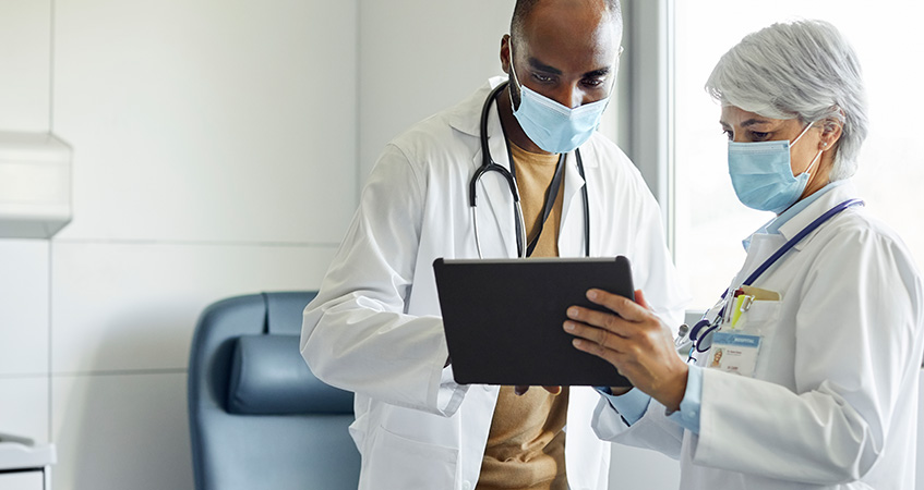 Two doctors wearing masks and white coats stand in a medical office, looking at a tablet together. They collaborate on patient information, using data-driven tools to enhance diagnosis and care decisions.
