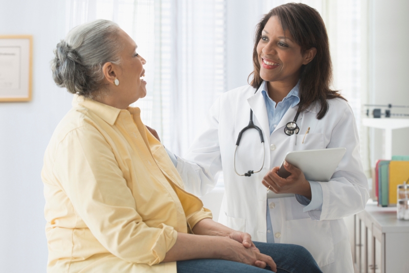 A smiling female doctor with a stethoscope and tablet discusses RCM challenges facing mental health providers with an older woman in a yellow shirt, seated on an exam table in a bright medical office.