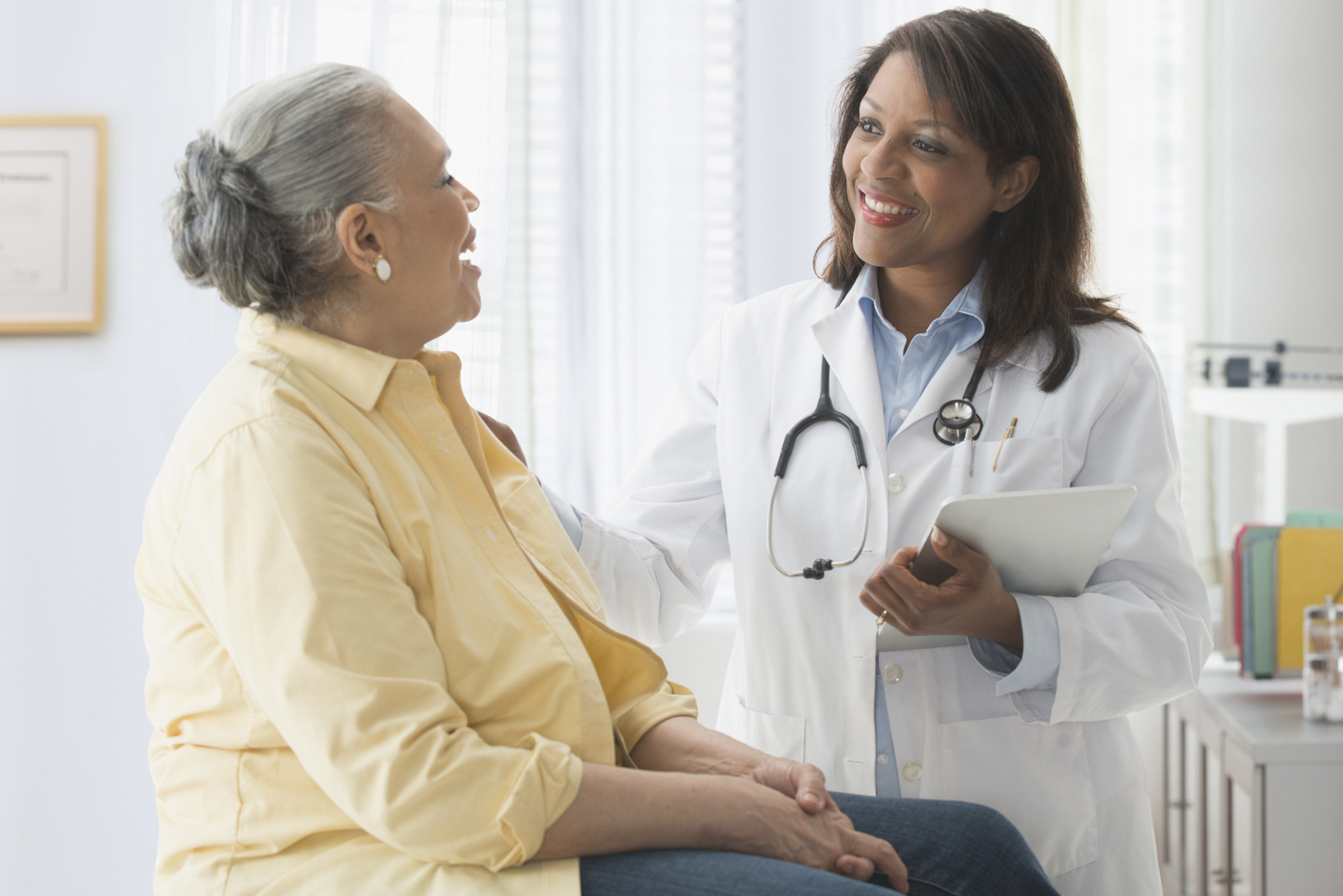 A female doctor with a stethoscope smiles and talks with an older woman sitting on an exam table in a bright medical office, discussing RCM challenges facing mental health providers as she reviews notes on her tablet and gently rests her hand on the patient’s shoulder.