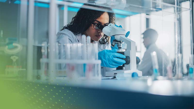 A scientist wearing gloves and a lab coat examines a sample through a microscope in a laboratory, surrounded by glass test tubes and equipment designed to boost lab performance.