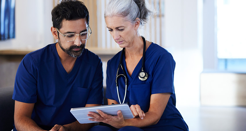 Two healthcare professionals in blue scrubs, one older woman with gray hair and a stethoscope, and a younger man with glasses and a beard, sit together looking at a tablet screen, appearing focused on ways to improve revenue cycle KPIs.