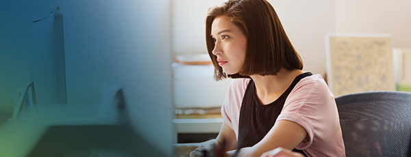 A young woman with short brown hair sits at a desk, focused on a computer screen. She is wearing a light pink shirt and works in a modern office, collaborating with a new clearinghouse partner under soft lighting.