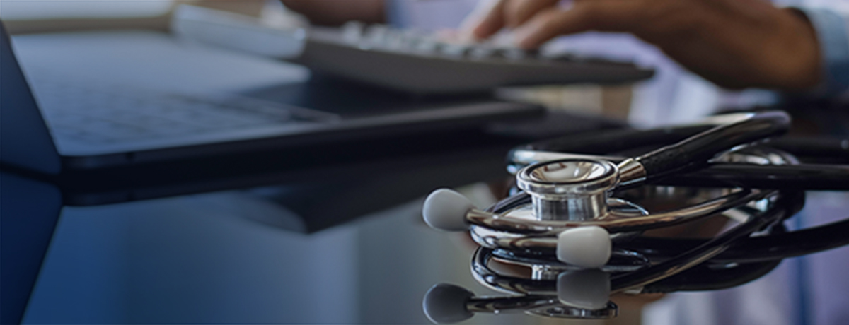 A stethoscope rests on a glossy desk near a laptop, while a person types on a keyboard in the background, reflecting efforts to enhance billing services in a modern medical office.