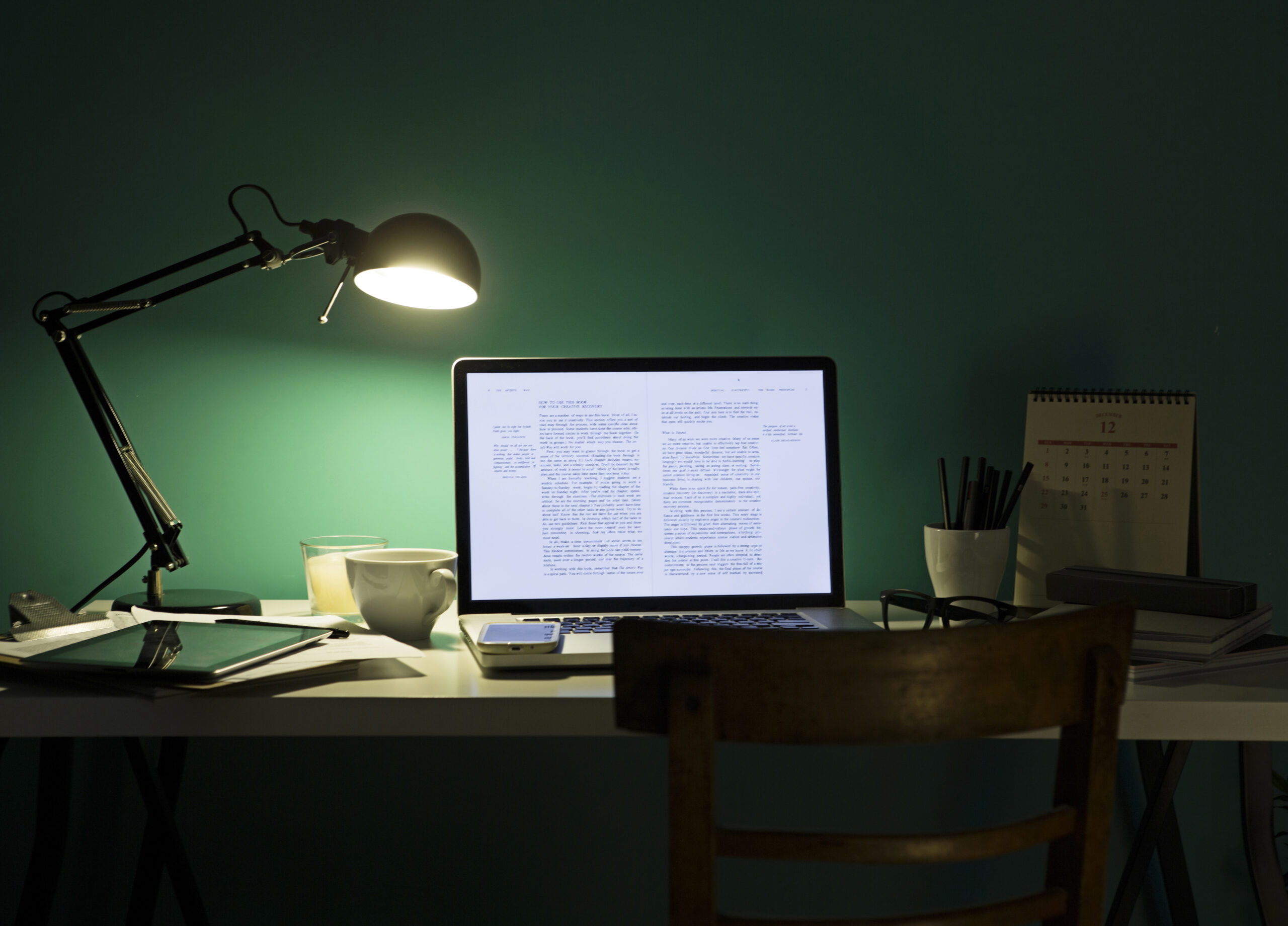 A desk for research and publications, with a laptop displaying text, a desk lamp switched on, a cup, tablet, phone, glasses, and calendar—all set against a teal wall with a wooden chair in front.