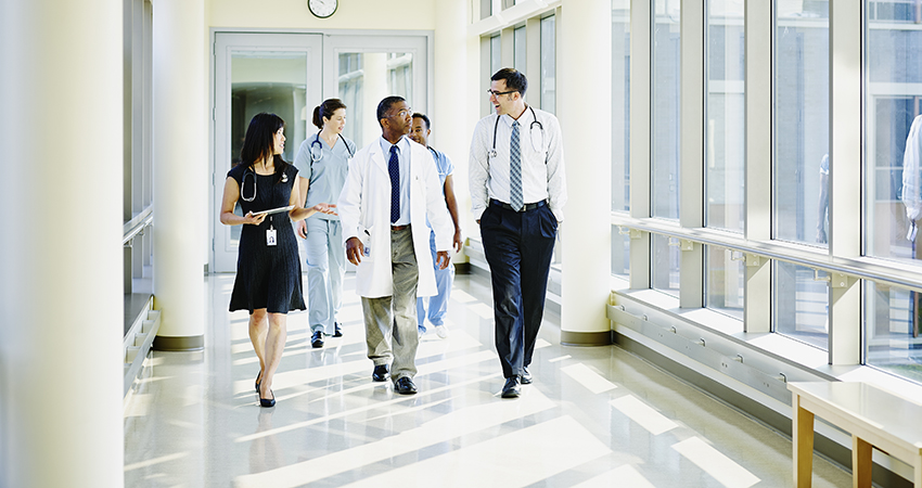 A group of medical professionals, including doctors and nurses, walk together down a bright hospital corridor, engaged in conversation. Large windows line the hallway, letting in natural light.