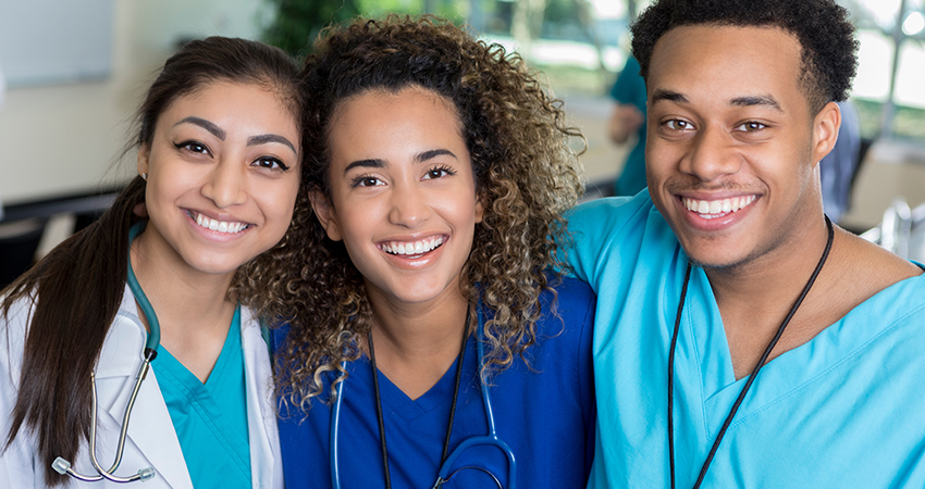 Three young healthcare professionals, two women and one man, wearing scrubs and stethoscopes, smile and pose together in a bright medical setting.