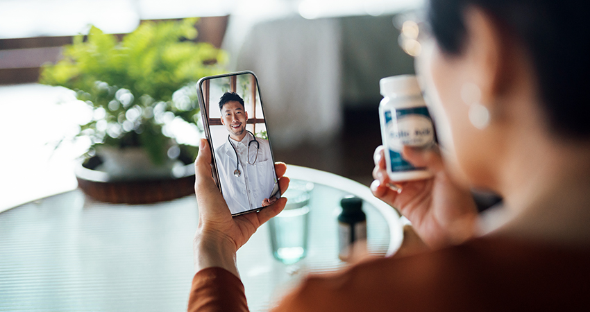 A person uses telehealth technology, holding a smartphone with a video call to a doctor while holding a medication bottle. A green plant and a glass of water sit on the table in front of them.