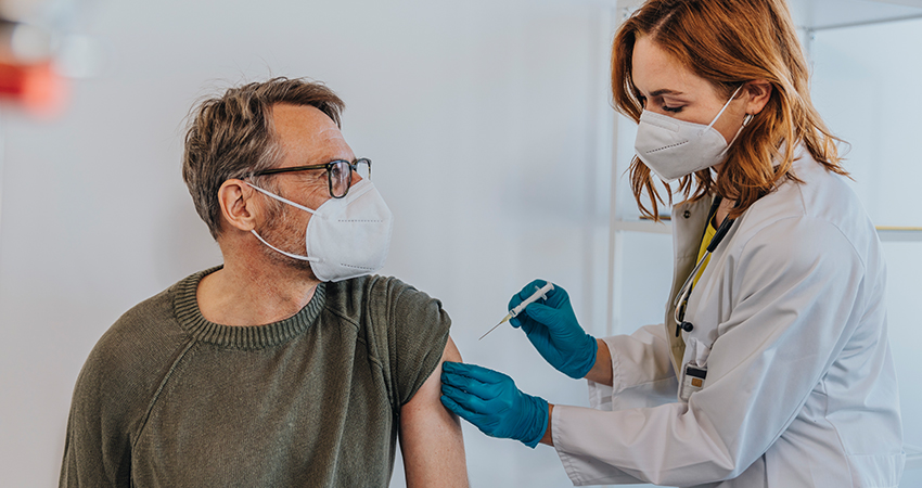 A healthcare worker wearing a mask and gloves administers a vaccine injection to a masked man’s upper arm in a clinical setting, promoting vaccine adherence.