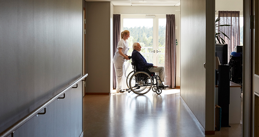A nurse stands behind an elderly man in a wheelchair as they gaze out large windows at the end of a hallway, reflecting the calm atmosphere and compassionate care found in today’s skilled nursing facility trends.