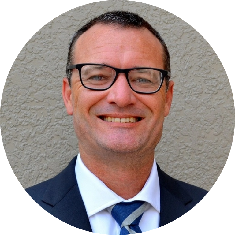 A man wearing glasses, a dark suit, and a blue-and-white striped tie smiles in front of a textured beige wall.