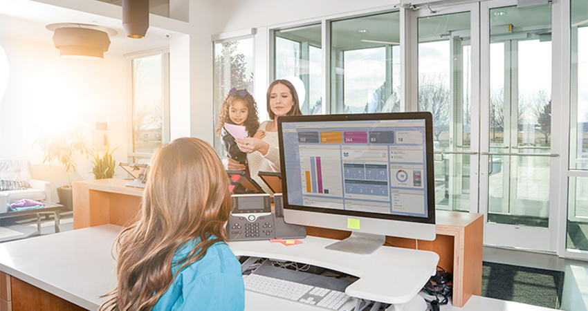 A receptionist sits at a desk with a computer displaying charts that help automate healthcare claims, while a woman and a young girl stand on the other side of the counter, handing over a document in a bright, modern office lobby.