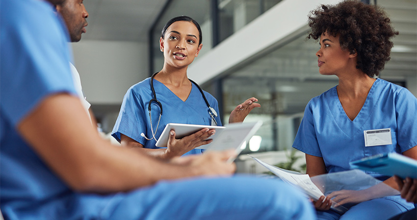 Three healthcare professionals in blue scrubs sit together indoors, holding papers and a tablet, discussing healthcare staffing needs in a modern medical setting.