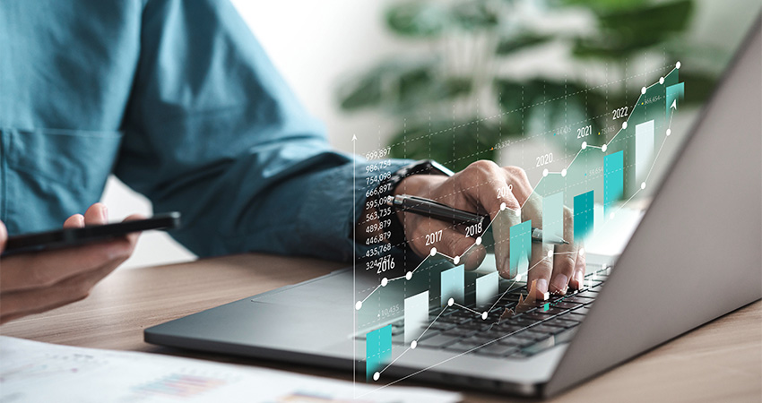 A person works on a laptop while holding a smartphone, with a digital graph showing rising bars and data points from 2016 to 2022 overlaid in the foreground, symbolizing financial growth or revenue cycle management analytics.