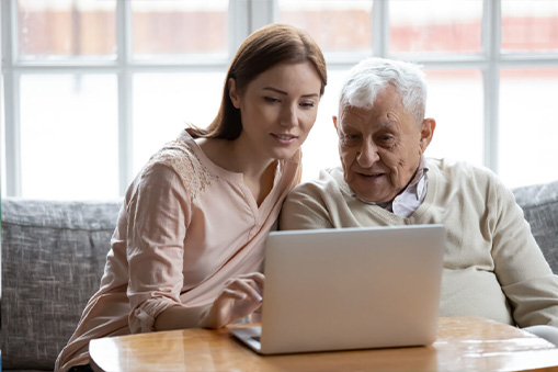 A young woman and an elderly man sit together on a couch, smiling at a laptop screen as they explore Real World Data, with a bright window in the background.