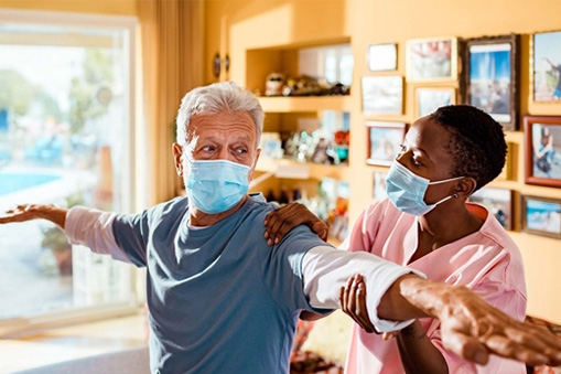 An older man and a healthcare worker, both wearing face masks, practice stretching exercises together in a brightly lit living room filled with photos and decorations, reflecting real world data on the benefits of at-home wellness routines.