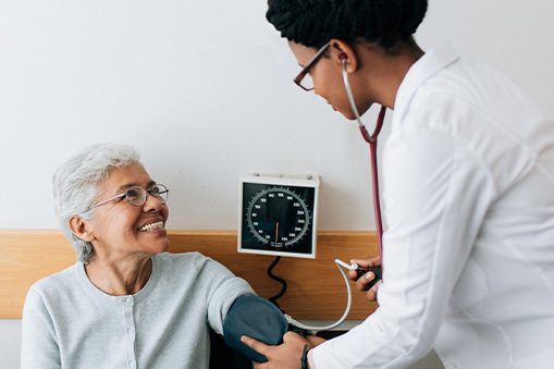 A doctor uses a blood pressure cuff to check an older woman's blood pressure, gathering real world data. The woman smiles at the doctor, who is wearing a white coat and using a stethoscope. A blood pressure monitor is visible on the wall.