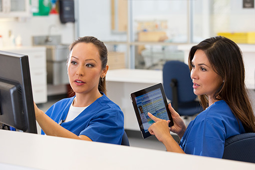 Two female healthcare workers in blue scrubs sit at a desk; one points at a computer monitor while the other holds a tablet displaying patient information. They appear to be discussing medical records and analyzing Real World Data.