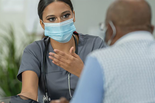 A female healthcare worker wearing a medical mask and scrubs speaks to an older male patient, explaining how Real World Data can impact his treatment, as she gestures with her hand in a clinical setting.