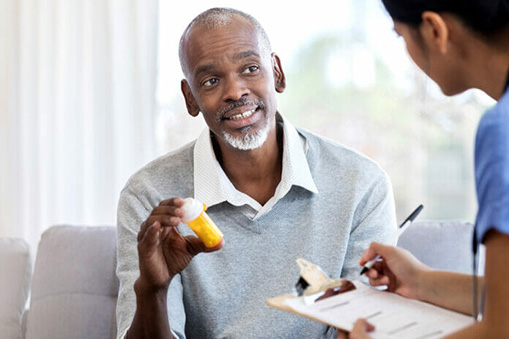 An older man sits on a couch, holding a prescription pill bottle and discussing his medication with a healthcare professional, who is taking notes—gathering real world data—in a comfortable home or clinic setting.