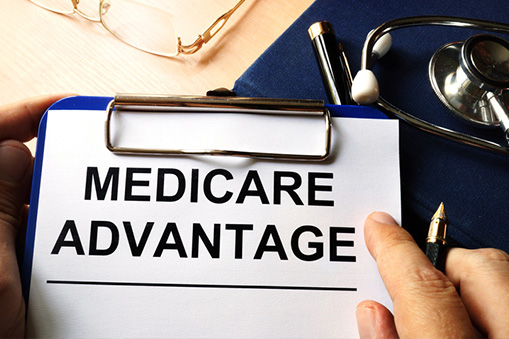A clipboard with a paper labeled Medicare Advantage held by a hand, alongside a stethoscope, glasses, and a blue folder on a desk—highlighting the importance of Real World Data in healthcare decision-making.