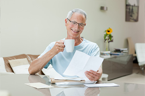 Older man with gray hair and glasses sits at a table, smiling while holding a white mug and reading real world data papers. An envelope and more documents are on the table in his bright living room.