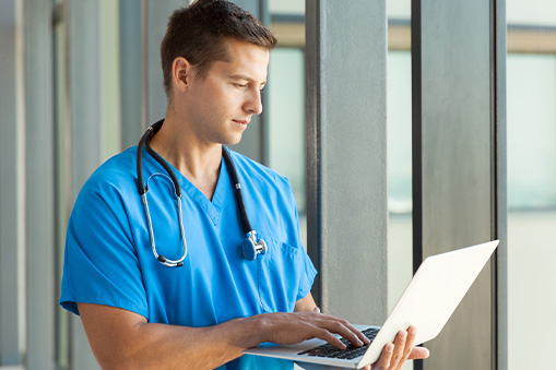 A male healthcare professional in blue scrubs with a stethoscope around his neck uses a laptop to review Real World Data while standing next to large windows.