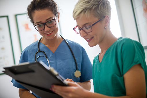 Two healthcare professionals wearing scrubs and glasses smile while reviewing Real World Data on a clipboard together in a medical office setting. One has a stethoscope around her neck.