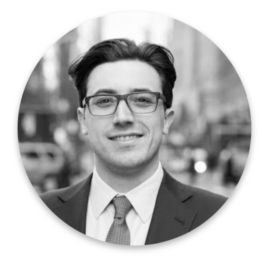 A man wearing glasses, a suit, and a tie is smiling at the camera. The background features a blurred urban street scene, emphasizing the subject. This black and white image, cropped into a circle, captures a professional in the realm of Real World Data.