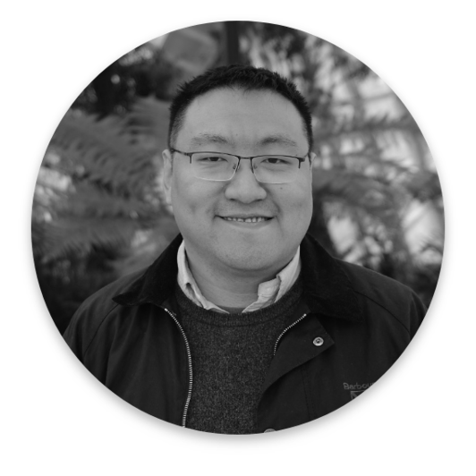 A man with short hair and glasses smiles at the camera. He is wearing a jacket over a sweater and shirt. The blurred foliage background evokes the nuanced perspective of Real World Data. The image is in black and white with a circular frame.