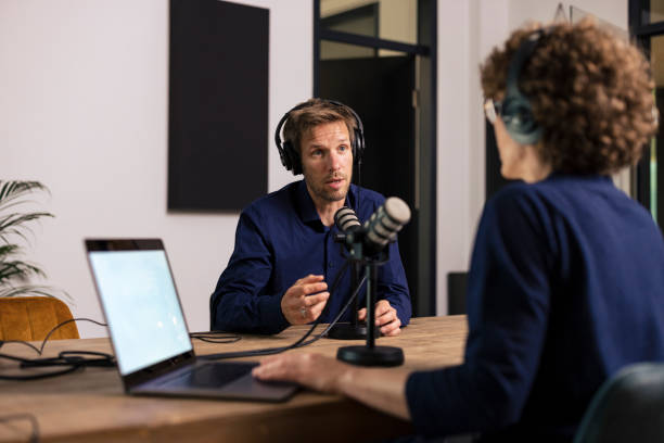Two people wearing headphones sit at a table with microphones and a laptop, discussing Real World Data as they record a podcast or interview in a modern studio. One person is speaking while the other listens attentively.