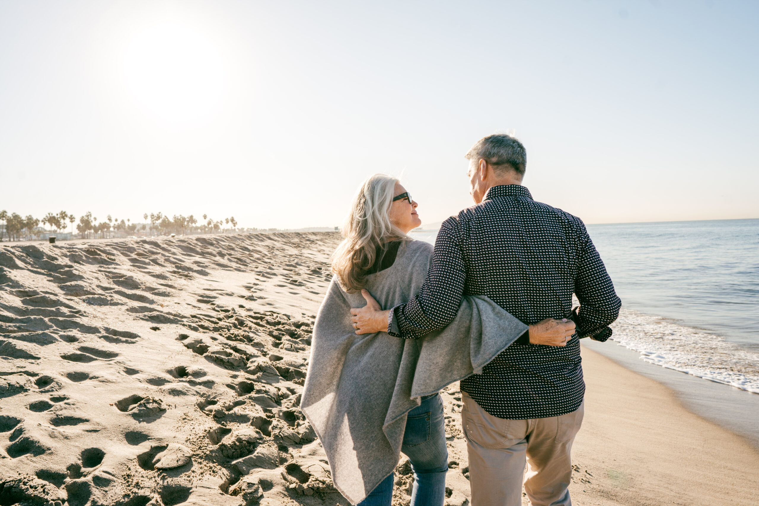 An older couple walks arm in arm along a sandy beach at sunset, with gentle waves and palm trees in the background—capturing a serene moment that reflects genuine connection, much like insights drawn from Real World Data.
