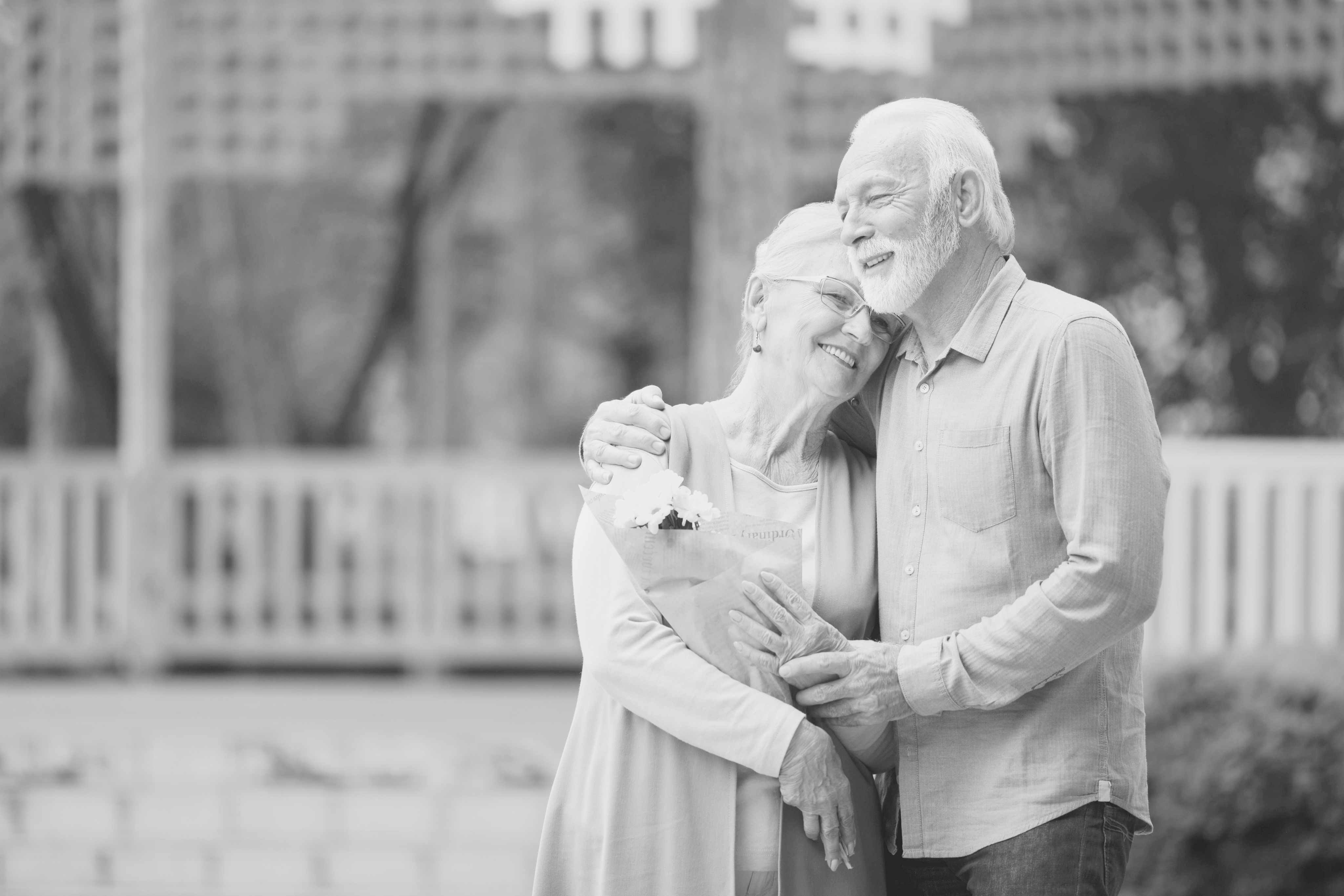 An elderly couple embraces and smiles outdoors, reflecting real world data on happiness in later life. The woman holds a bouquet, resting her head on the man's chest. Both appear content against a softly blurred background of trees and a fence.