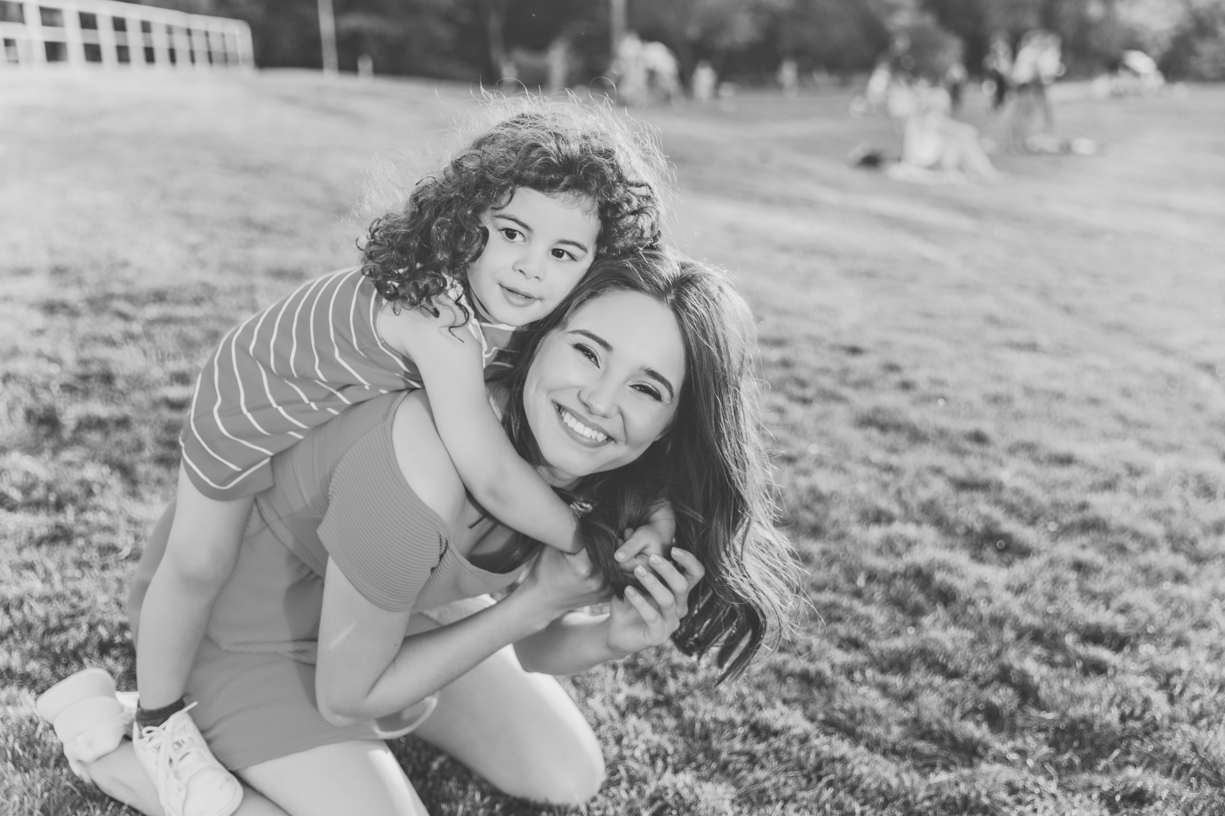 A woman kneels on grass, smiling widely, while a young girl with curly hair hugs her from behind. Capturing Real World Data, the pair appear happy outdoors in a sunny park with people in the background.