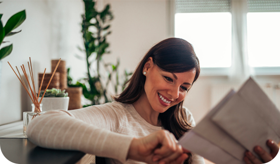 A smiling woman sits indoors and reads a letter, surrounded by houseplants and soft natural light from a window in the background, creating a serene scene that feels inspired by real world data.