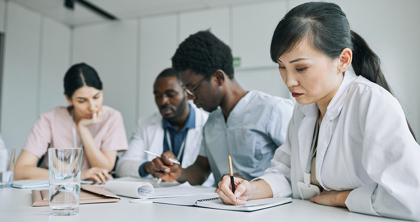 Four healthcare professionals sit at a table in a meeting room, focused on notes and documents related to regulatory compliance. Three are writing and one is reading, with glasses of water and papers spread out before them.