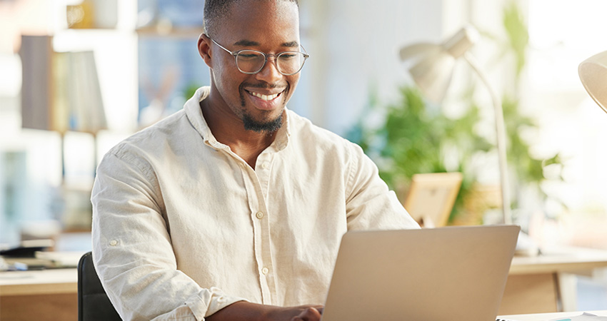 A man wearing glasses and a light-colored shirt smiles while working on a laptop with risk adjustment software at a bright, modern desk, surrounded by plants and office supplies.