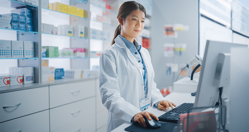 A pharmacist in a white lab coat stands at a pharmacy counter, using a computer. Shelves filled with medication boxes line the background, reflecting the bright, modern setting and hinting at the future of pharmacy.