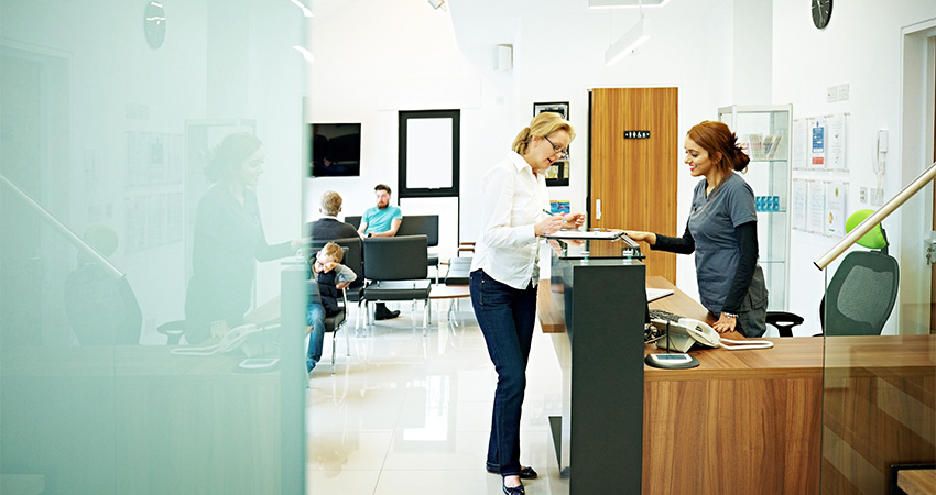 A woman stands at a reception desk speaking to a receptionist about patient demographic data verification in a modern, bright waiting room, with several people sitting and talking in the background.
