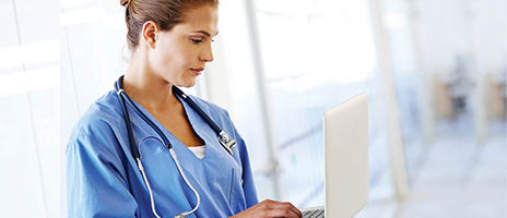 A healthcare professional in blue scrubs with a stethoscope around their neck is working on a laptop in a bright, clinical setting.