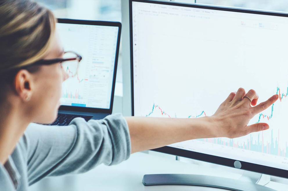 A person wearing glasses points at a rising stock chart displayed on a computer monitor, with another laptop in the background showcasing real world data and financial information.