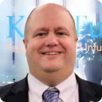 A smiling bald man in a suit and tie stands in front of a blurred blue and white background with indistinct text and shapes.