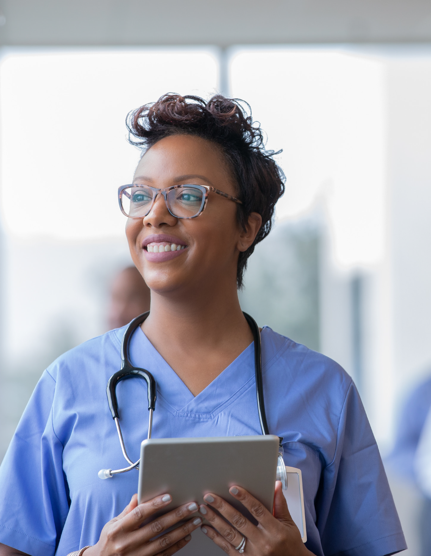 A healthcare professional in blue scrubs and glasses smiles while holding a tablet. She has a stethoscope around her neck and stands in a bright, modern medical setting.