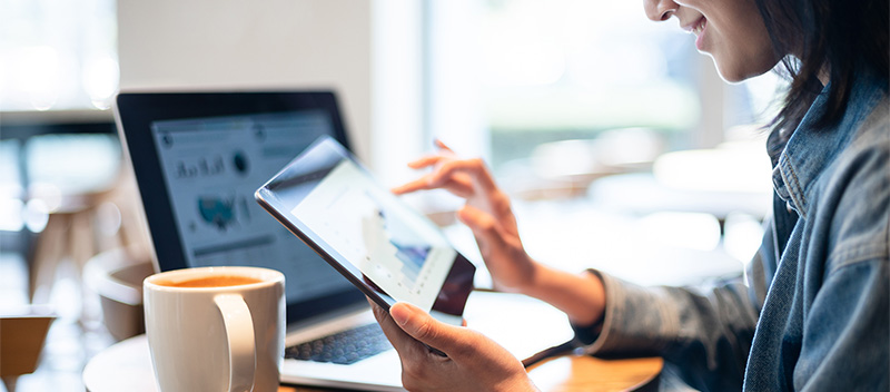 A person sitting at a café table uses a tablet with one hand, while a laptop displaying SDOH analytics and a cup of coffee are on the table. The person is smiling and appears to be working or studying.