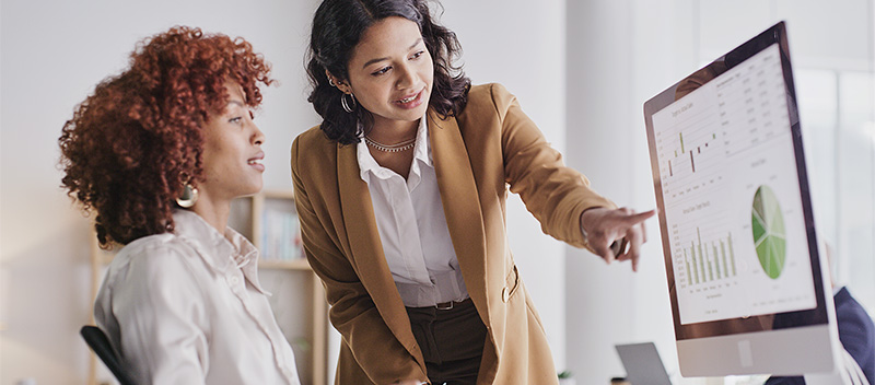 Two women in business attire review HEOR data on a computer monitor displaying graphs and charts. One is seated, while the other points at the screen, indicating a collaborative discussion in a modern office setting.