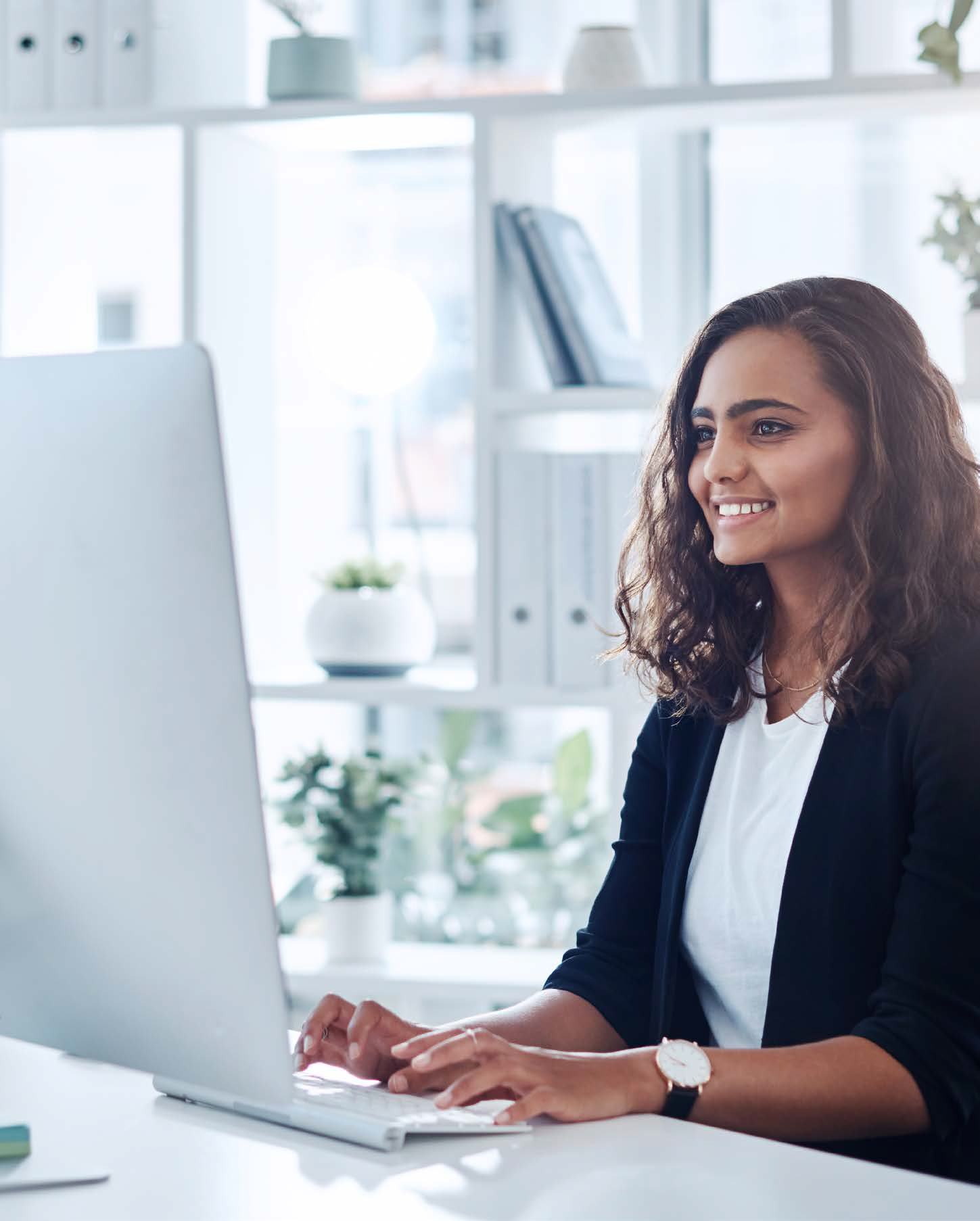 A woman with curly hair, wearing a black blazer and white shirt, is smiling while typing on a computer at a bright, modern office desk with plants and shelves in the background.