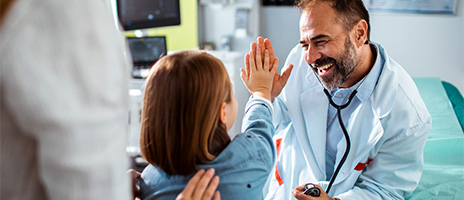 A smiling doctor in a white coat gives a high five to a young patient in a medical office, while another adult stands nearby, gently touching the childs shoulder.
