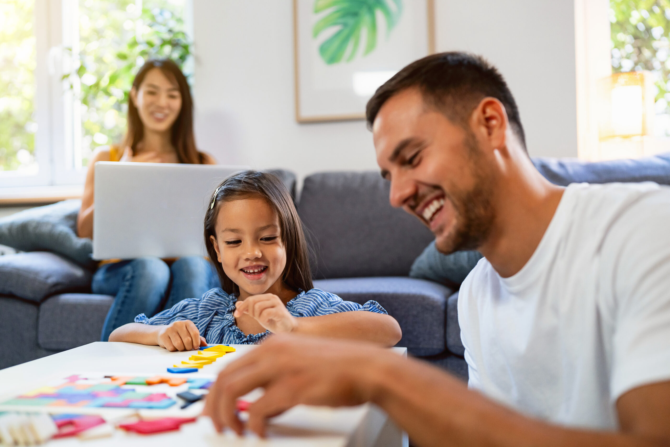A man and young girl smile while playing with colorful puzzle pieces at a table. A woman sits on a couch in the background, using a laptop. The setting is a bright, cozy living room.