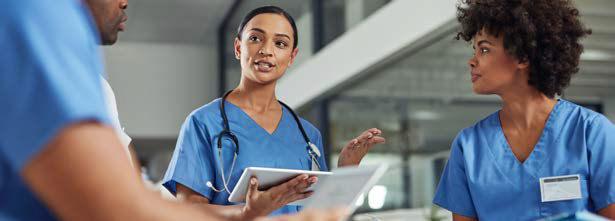 Three healthcare professionals in blue scrubs are having a discussion indoors. One person is holding a digital tablet, and all are engaged and focused, suggesting a collaborative work environment.