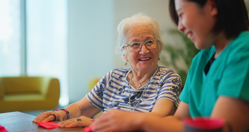 An elderly woman with white hair and glasses smiles while sitting at a table with a younger woman in green scrubs. They appear to be engaging in a craft at AAPACN 2024 in a bright, cheerful room.